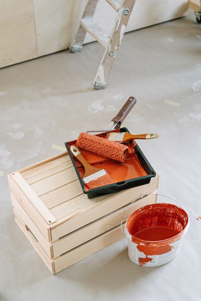 pexels photo 5799053 A close-up of home renovation tools including paint roller and red paint bucket on a wooden crate.